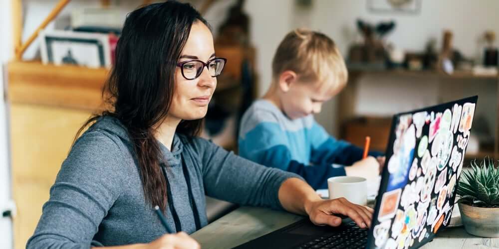 Mother working on laptop next to son coloring, sitting at kitchen table